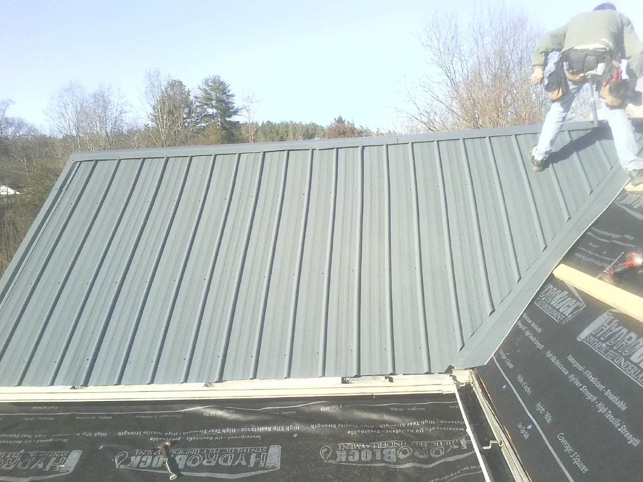 Crew installing dark green standing seam panels over synthetic underlayment in Petersham