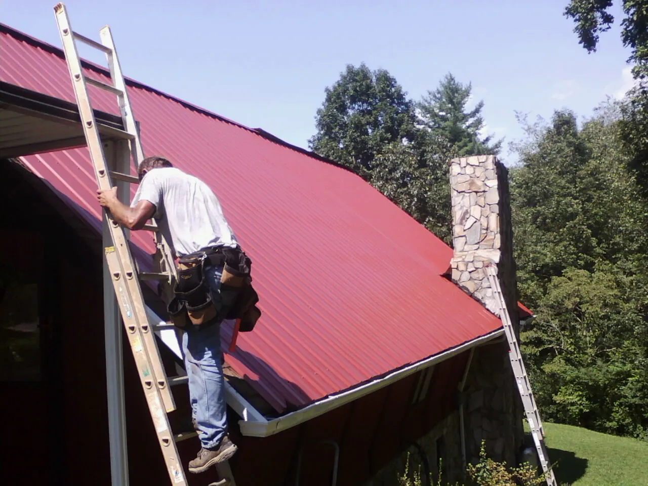 Roofer on a ladder beside a red metal roof and stone chimney — Soffit & Fascia Repair by Roofing Brothers in Petersham